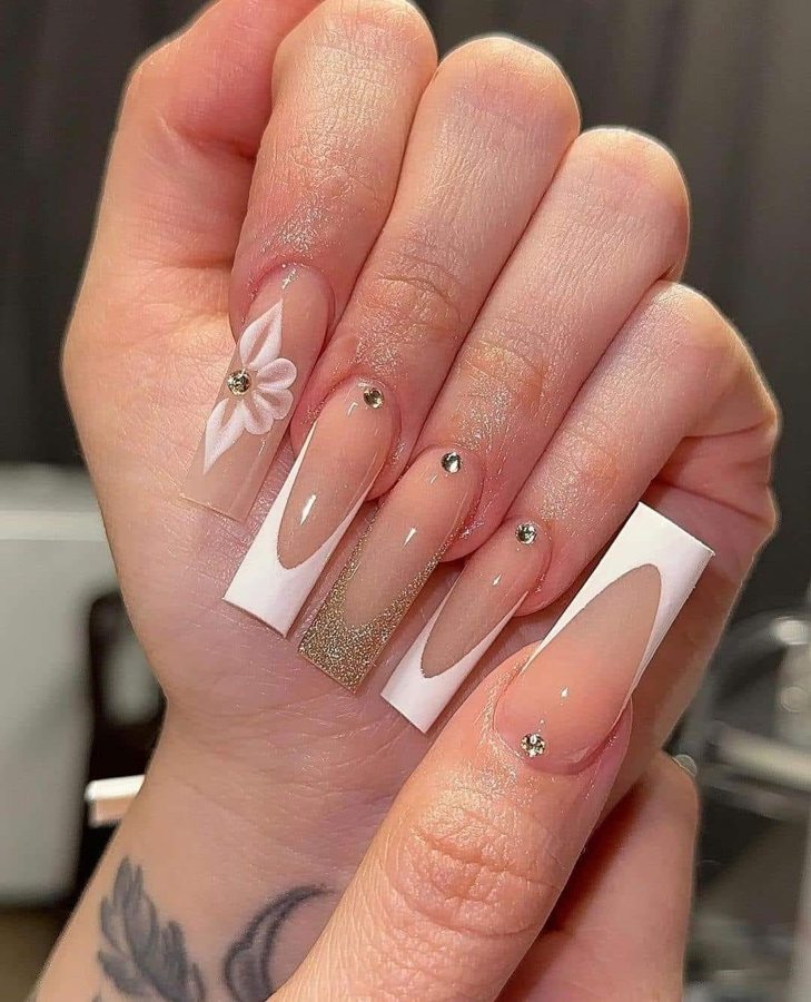 Long nude nails with white French tips, gold glitter, rhinestones and a 3D white flower